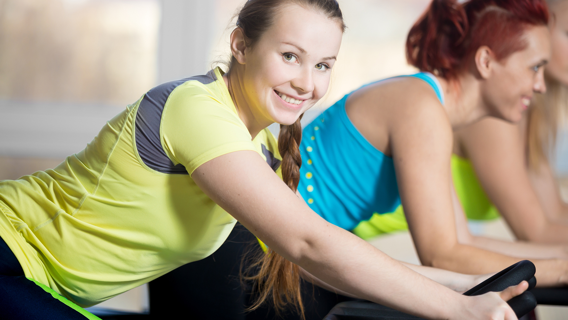 Fitness practice, group of beautiful cheerful females cycling in sports club, doing cardio exercises for slimming, warming up during sport lesson in class, focus on smiling woman with braid