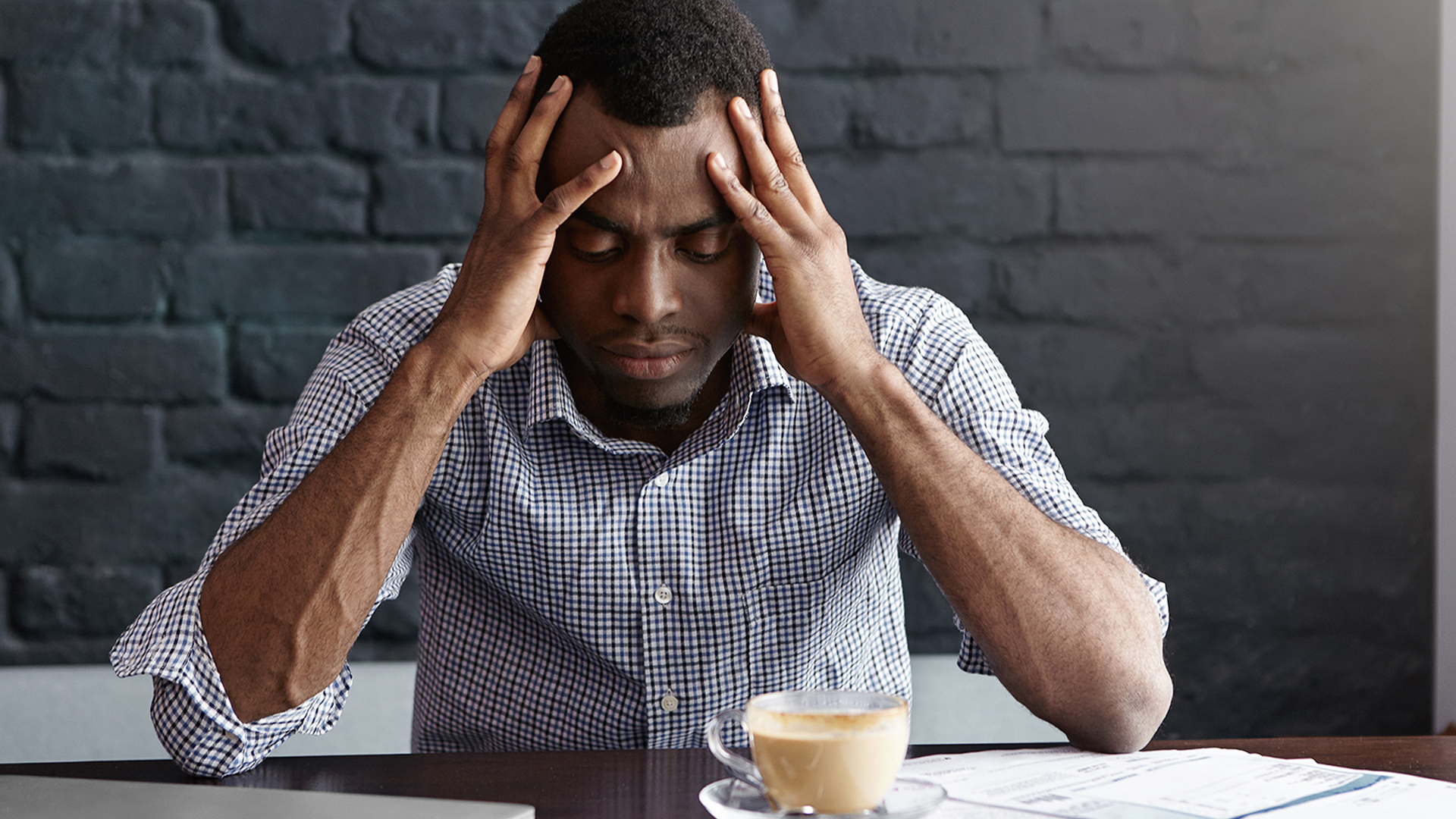 Frustrated young African-American businessman having bad headache, squeezing his temples, feeling stressed at work, sitting at desk with generic laptop computer, documents, mug and mobile phone
