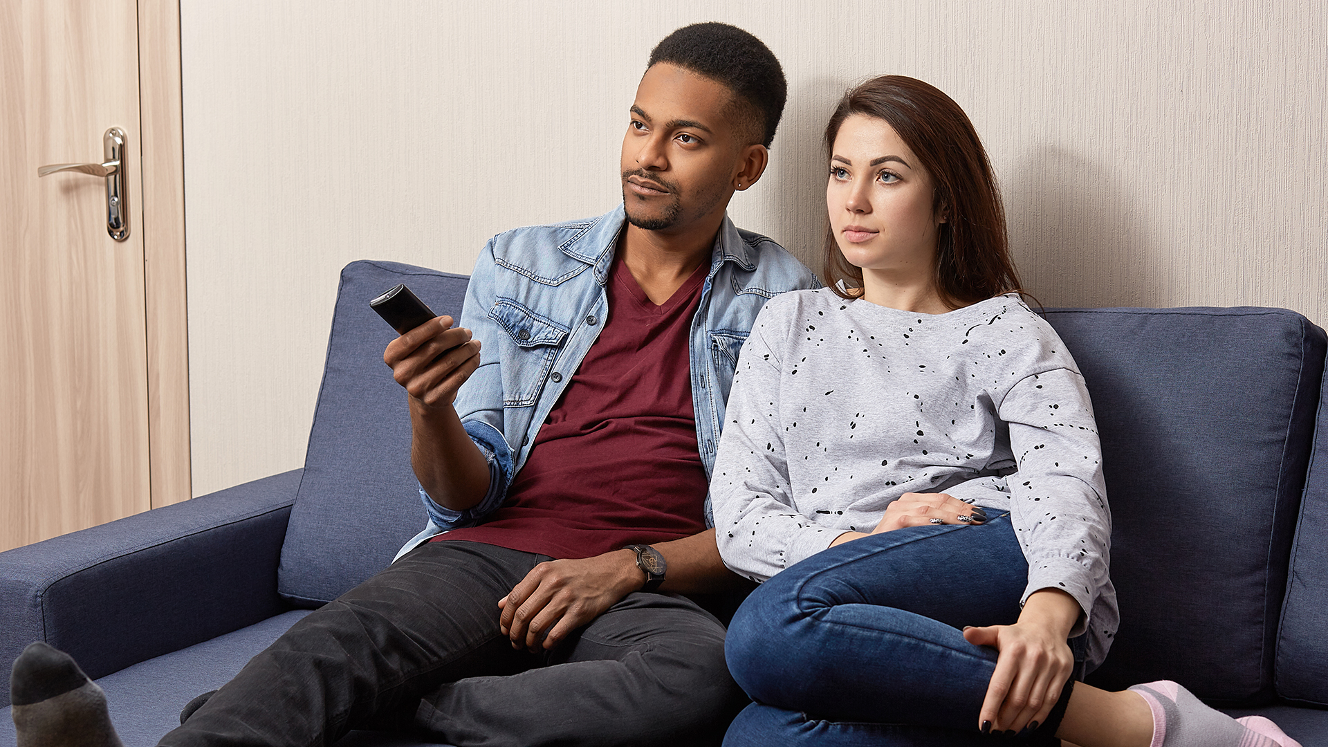 Indoor shot of multiethnic couple watch television at home on comfortable sofa. Black man holds remote control, switches on TV, sits near his young wife, have lazy day together, enjoy serial or movie