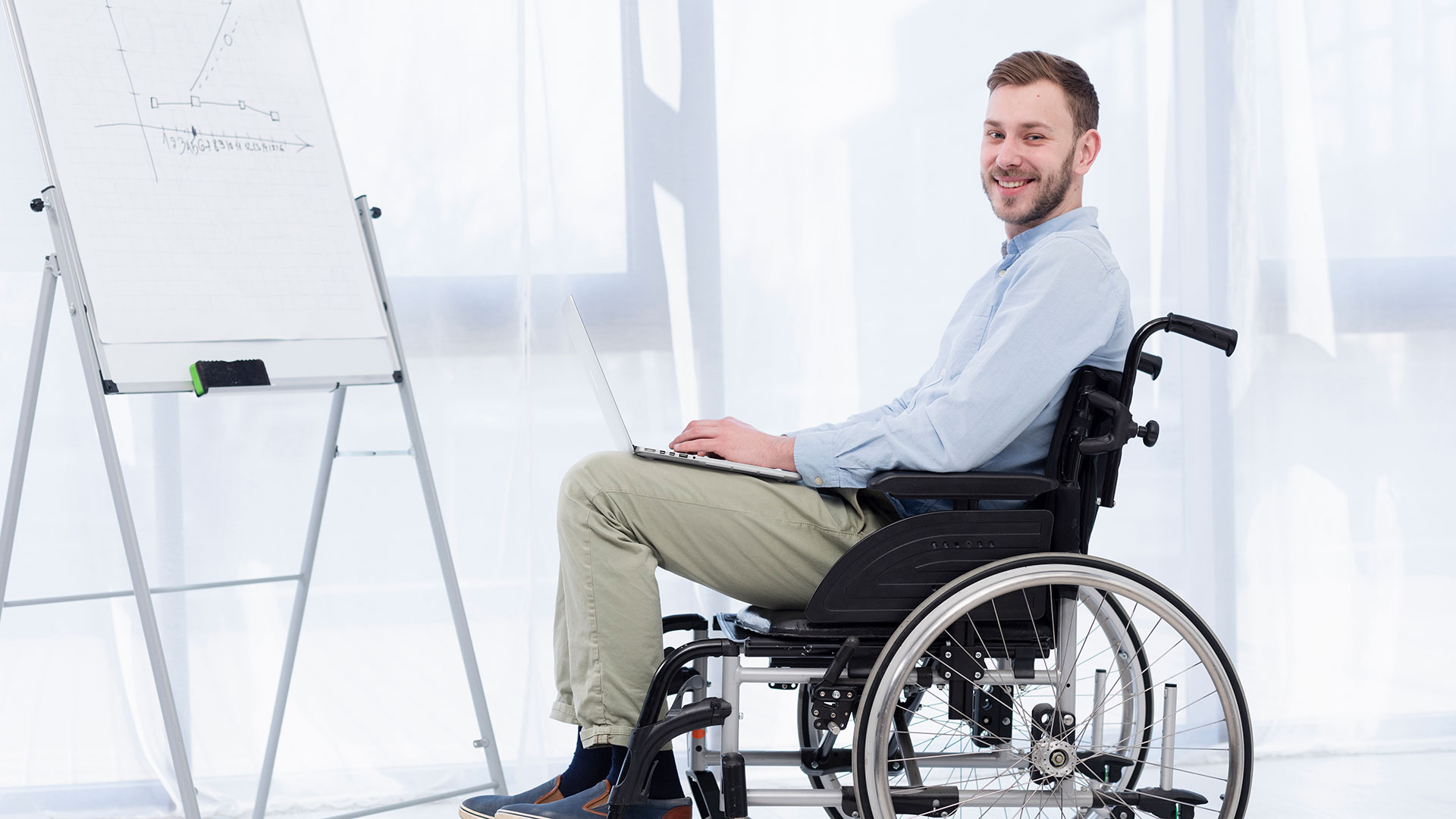 Disabled white man in wheelchair with laptop on his lap smiles at the camera.