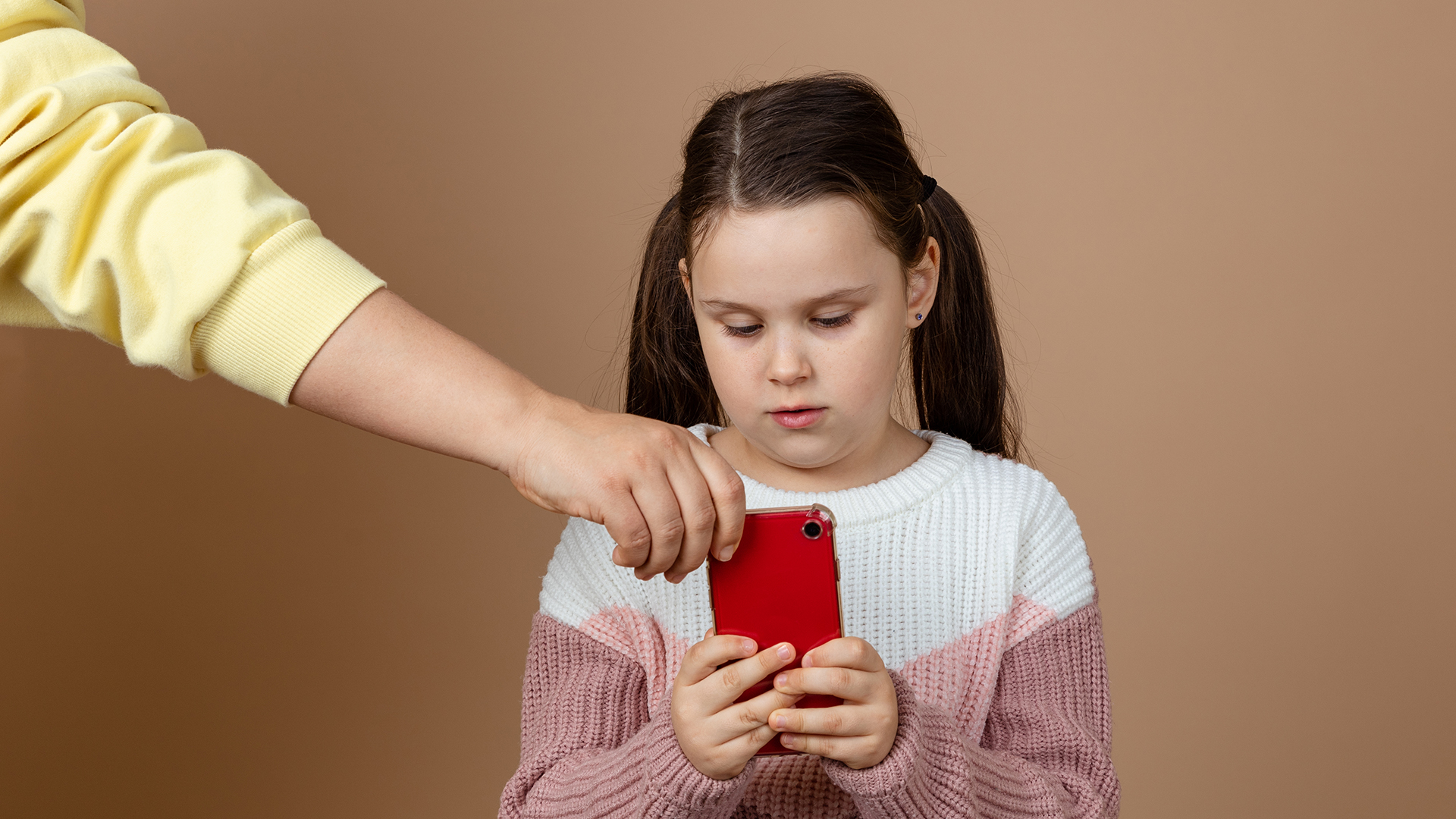 Portrait of parent taking smartphone away from girls hands, beige background. Little daughter hold tight phone and resist. Concept of prevention of telephone addiction in children, nomophobia