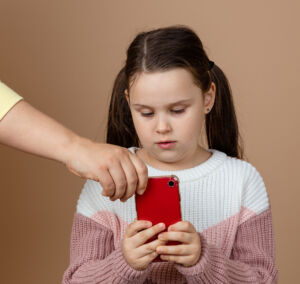 Portrait of parent taking smartphone away from girls hands, beige background. Little daughter hold tight phone and resist. Concept of prevention of telephone addiction in children, nomophobia.