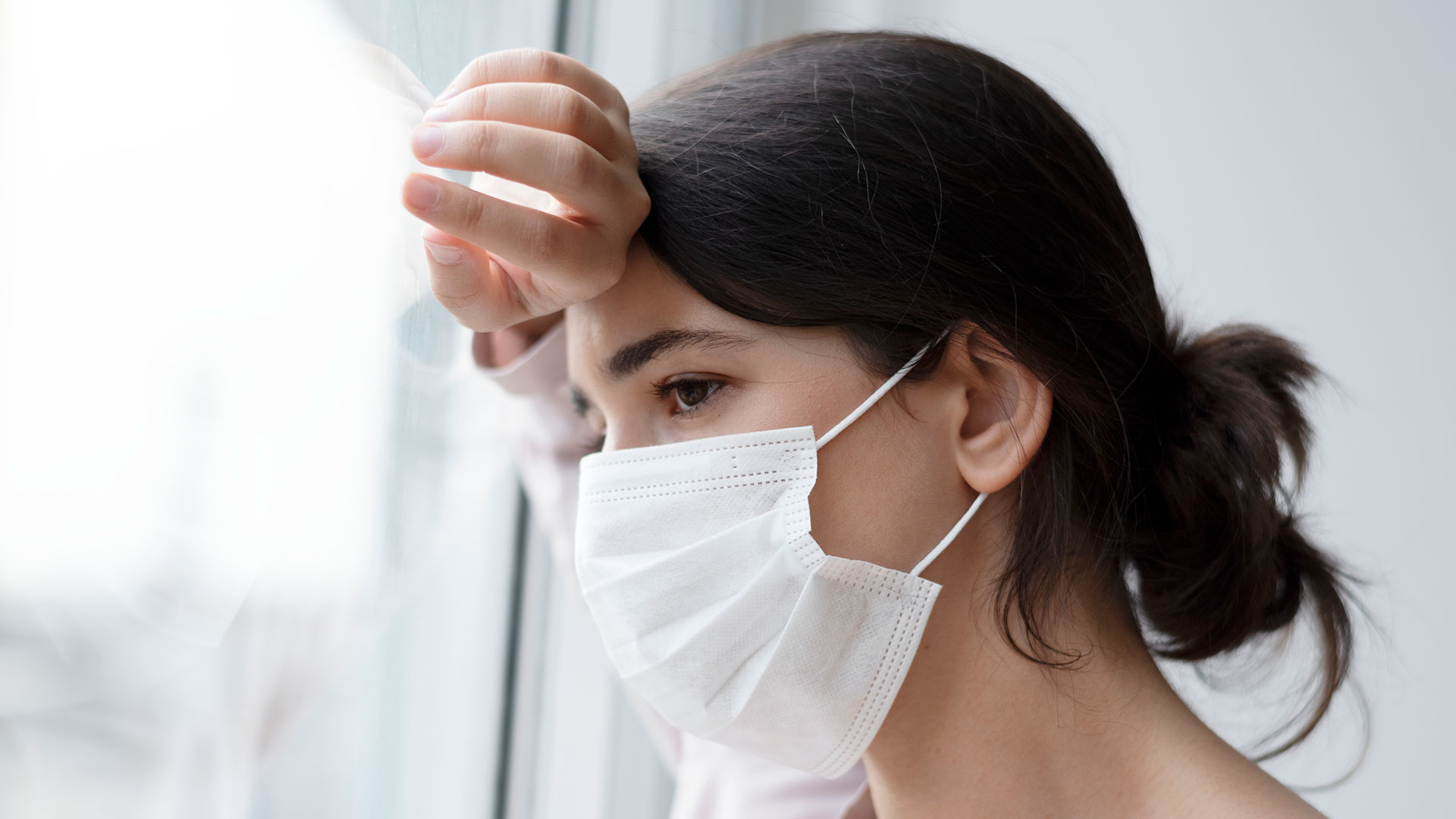 young woman with dark hair wearing a face mask, looks longingly out the window.